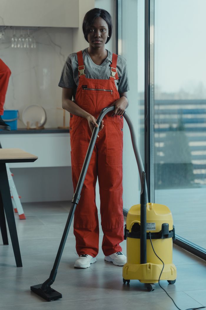 An adult woman wearing red coveralls is using a vacuum cleaner in a contemporary indoor space, concentrating on cleaning work.